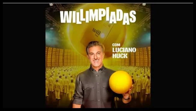 Luciano Huck segurando uma bola amarela em frente a uma plateia vestida de amarelo, com o logo da promoção Willimpíadas do Will Bank 2025 ao fundo.