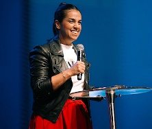 Pastora Camila Barros ministrando em um evento cristão, com microfone na mão, em um palco iluminado, diante de uma plateia atenta.